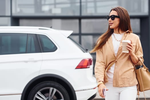 Outdoors against modern building. Fashionable beautiful young woman and her modern automobile.