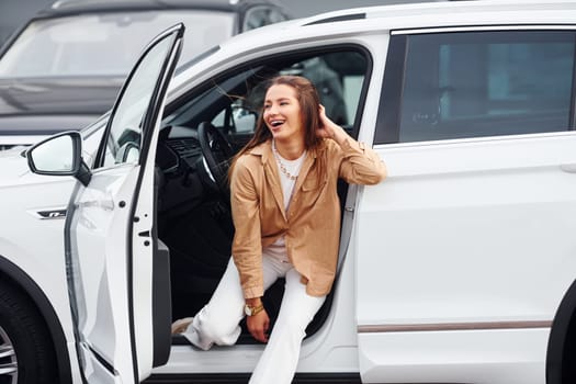 Outdoors against modern building. Fashionable beautiful young woman and her modern automobile.