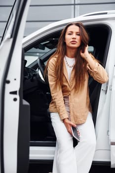 Outdoors against modern building. Fashionable beautiful young woman and her modern automobile.