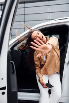 Outdoors against modern building. Fashionable beautiful young woman and her modern automobile.