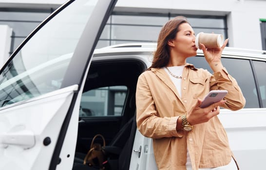 With coffee and phone. Fashionable beautiful young woman and her modern automobile.