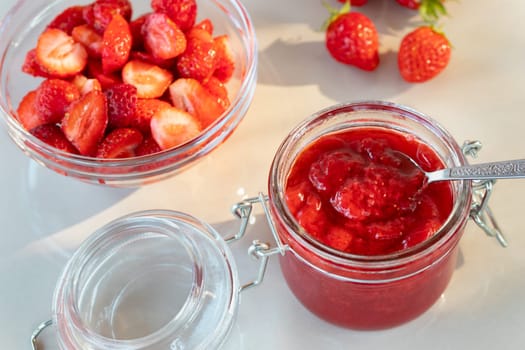 Glass jar with strawberry jam prepared for canning and fresh strawberries in a bowl on the table.