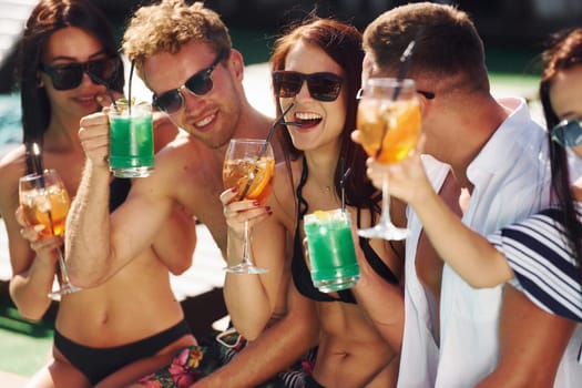 Fresh cocktails in hands. Group of young happy people have fun in swimming pool at daytime.