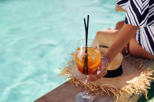 Young woman sits near swimming pool at daytime with cocktail.