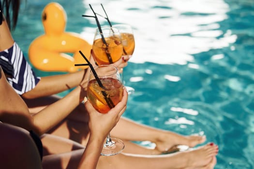 Fresh cocktails in hands. Group of young happy people have fun in swimming pool at daytime.