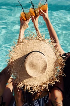 Fresh cocktails in hands. Group of young happy people have fun in swimming pool at daytime.