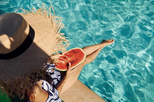 Young woman sits near swimming pool at daytime with watermelon.