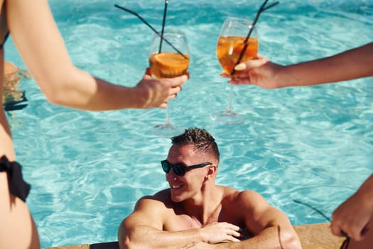 Fresh cocktails in hands. Group of young happy people have fun in swimming pool at daytime.