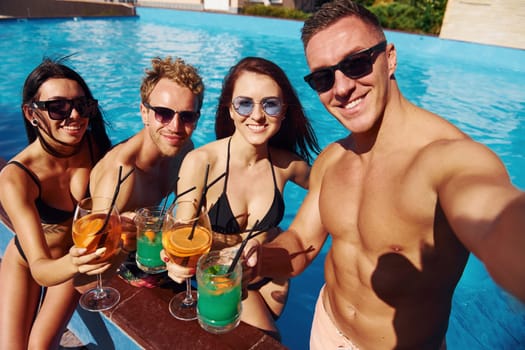 Making selfie. Group of young happy people have fun in swimming pool at daytime.