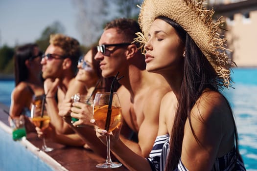 Talking and drinking. Group of young happy people have fun in swimming pool at daytime.