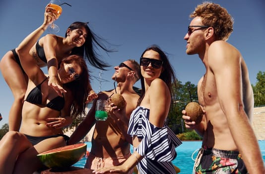 Holding watermelon. Group of young happy people have fun in swimming pool at daytime.