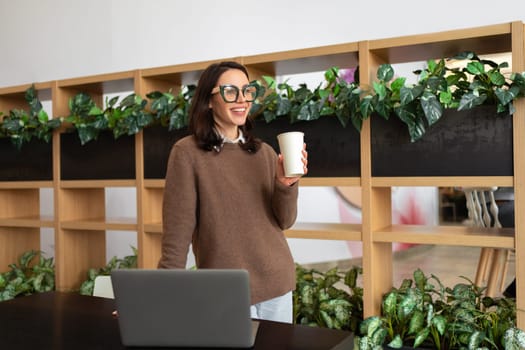 Happy European Female Entrepreneur Standing Near Desk With Laptop Computer Posing Smiling In Modern Office Indoor. Freelance Career And Successful Entrepreneurship Concept