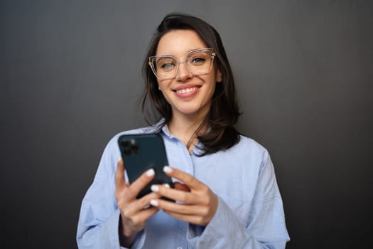 Satisfied smiling woman in glasses with smartphone, types text message on mobile phone, enjoys online communication, wears denim shirt, isolated on black studio background looking at camera