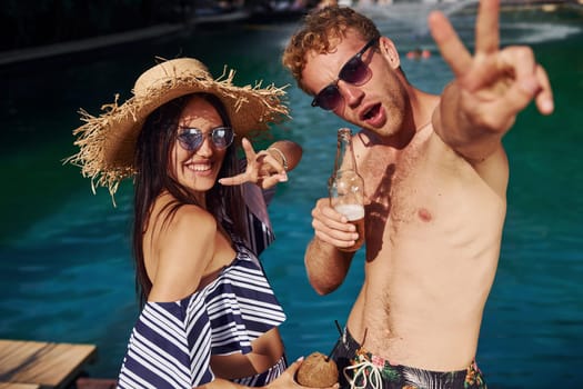Holding drinks in hands. Cheerful couple or friends together in swimming pool at vacation.