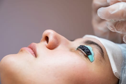 Close-up portrait of a woman on eyelash lamination procedure. The master applies tint to the eyelashes