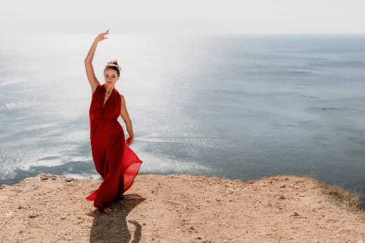 Side view a Young beautiful sensual woman in a red long dress posing on a rock high above the sea during sunrise. Girl on the nature on blue sky background. Fashion photo.