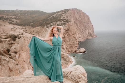 Side view a Young beautiful sensual woman in a mint long dress posing on a volcanic rock high above the sea during sunset. Girl on the nature on overcast sky background. Fashion photo