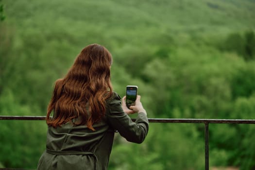 a woman in a raincoat takes pictures on the phone of a beautiful view of the forest. Travel, technology, mobile photography. High quality photo