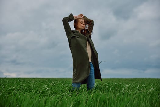 a woman in a long coat stands in a field against a cloudy sky with her back to the wind with her hands raised behind her head. High quality photo