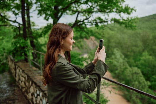 a woman in a raincoat takes pictures on the phone of a beautiful view of the forest. Travel, technology, mobile photography. High quality photo