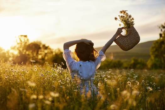 silhouette of a woman in a light dress sits in a chamomile field at sunset and holds a wicker basket of flowers raised in her hand. High quality photo