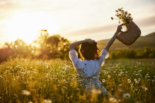 silhouette of a woman in a light dress sits in a chamomile field at sunset and holds a wicker basket of flowers raised in her hand. High quality photo