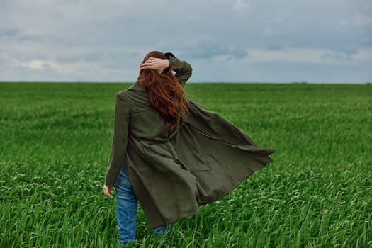 a red-haired woman stands with her back to the camera in a long coat, holding her hand with her hair flying in the wind, standing in a green field in cloudy weather. High quality photo