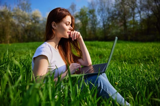 Young Caucasian woman using Laptop on nature, sitting in the grass in the park. High quality photo