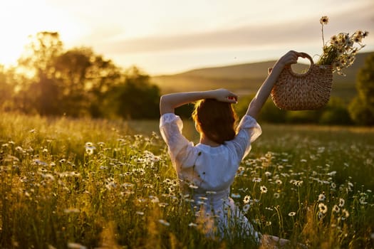 silhouette of a woman in a light dress sits in a chamomile field at sunset and holds a wicker basket of flowers raised in her hand. High quality photo