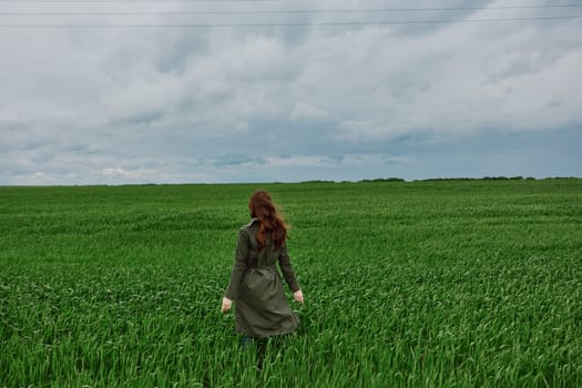 a red-haired woman in a long raincoat stands in a green field in cloudy weather with her back to the camera enjoying nature. High quality photo