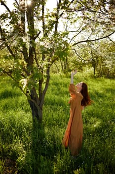 a beautiful, joyful woman stands in a long orange dress near a tree blooming with white flowers during sunset, illuminated from the back and joyfully raises her hands up. High quality photo