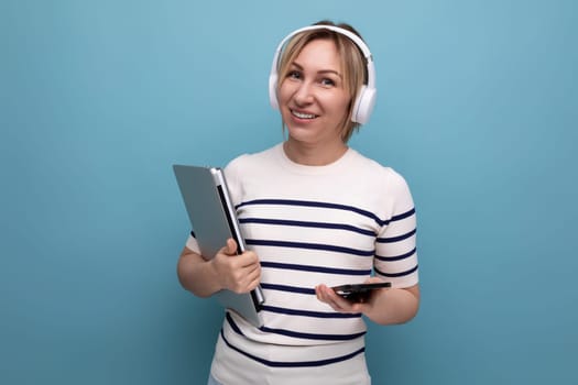 portrait of cute smiling blond young female student with big wireless headphones and laptop on blue background with copy space.