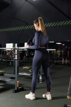 One adult caucasian woman female athlete putting weight plate on the barbel at gym. wearing blue leggins and long sleeve top