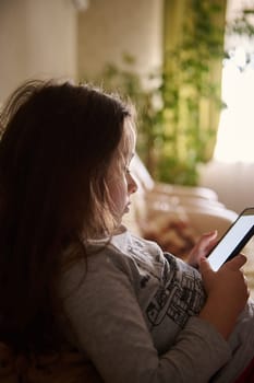 Caucasian beautiful cute little girl sitting on sofa and holding smartphone at home room, playing online virtual games and watching online videos. Side view. Children addiction. Spending time alone.