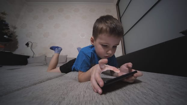The boy plays on his smartphone while lying on the couch at home