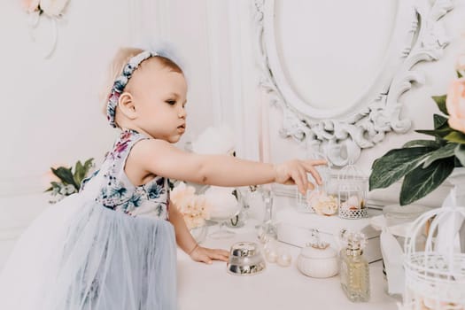 Baby girl elegant dress. A one-year-old girl in a puffy dress and a cute bow poses against the backdrop of a bright room with a dressing table and flowers