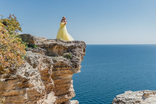 Woman in a yellow dress on the sea. Side view Young beautiful sensual woman in yellow long dress posing on a rock high above the sea at sunset. Girl in nature against the blue sky.