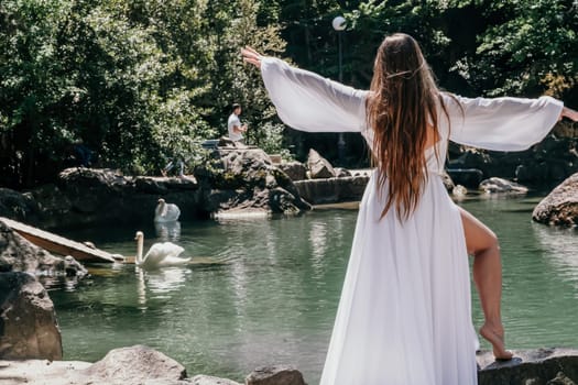 a beautiful woman in a long white dress looks into the distance at a beautiful lake with swans