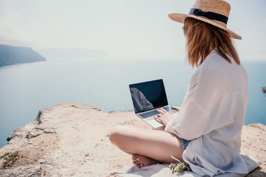 Successful business woman in yellow hat working on laptop by the sea. Pretty lady typing on computer at summer day outdoors. Freelance, travel and holidays concept.