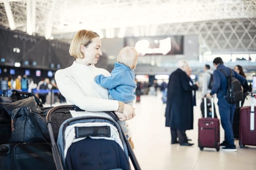 Motherat travelling with his infant baby boy child, walking, pushing baby stroller and luggage cart at airport terminal station. Travel with child concept