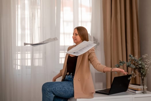 Professional woman sitting on desk in home office, positive female lawyer rejoices throwing papers up. She is wearing a beige jacket and jeans.