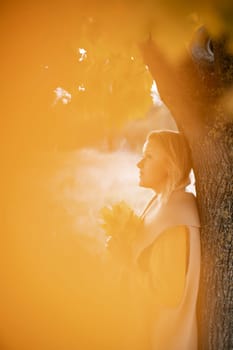 The blonde stands near the autumn tree. Thoughtful woman looks ahead, dressed in a yellow dress. Autumn content.