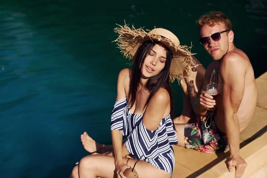 Holding drinks in hands. Cheerful couple or friends together in swimming pool at vacation.