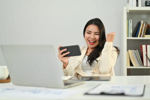 Overjoyed asian woman worker looking at mobile phone and celebrating success, excited by good news in email or message.