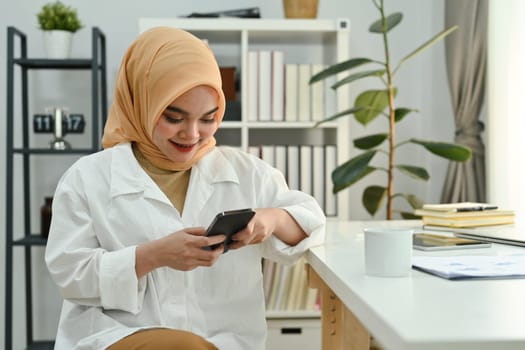 Smiling muslim woman sitting at the desk in modern office space and chatting online on mobile phone.
