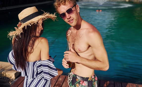 Holding drinks in hands. Cheerful couple or friends together in swimming pool at vacation.