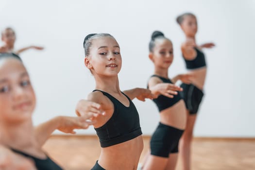 Standing and doing synchronised moves. Group of female kids practicing athletic exercises together indoors.