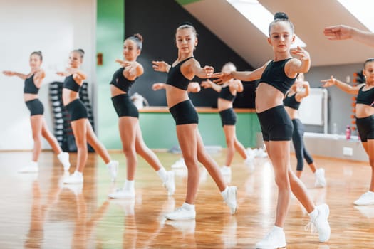 Standing and doing synchronised moves. Group of female kids practicing athletic exercises together indoors.