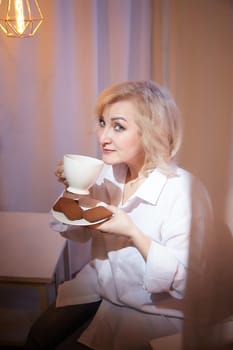 Adult mature woman of 40 or 60 years old with cup of coffee and chocolate cookies in casual white shirt and blue jeans. Calm cozy evening atmosphere with transparent curtains and soft warm lamp