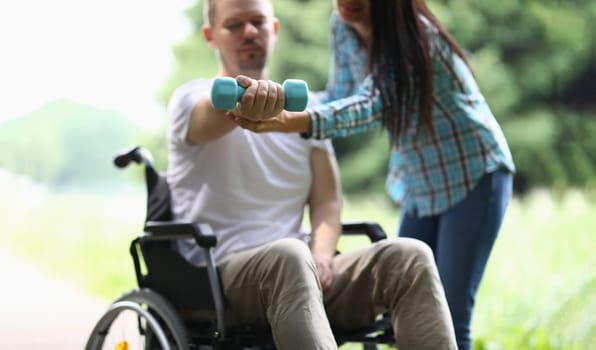 Female physiotherapist helps disabled man in wheelchair to lift weight of arm in park. Rehabilitation and exercises with dumbbells concept